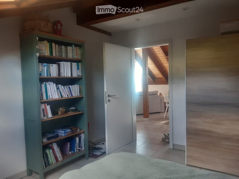 A cozy room with a green bookshelf filled with books, a white door leading to a living area with a sofa, a chair, and a small table with a lamp. The room has wooden beams on the ceiling and a soft, light-colored rug on the floor.
