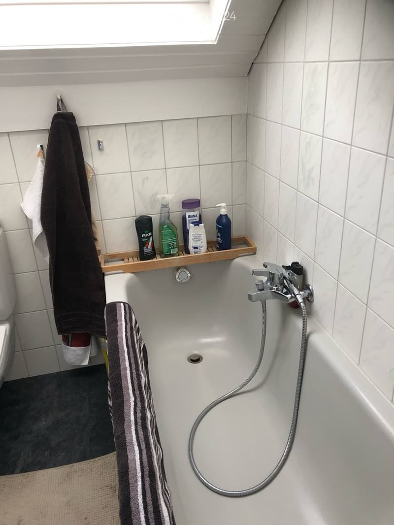 Bathroom with a white tiled wall, a bathtub with a handheld shower hose, a wooden tray holding several bottles, and towels hanging on the side.