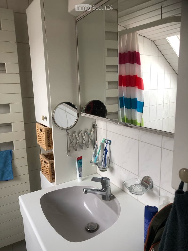 Modern bathroom with a white sink, chrome faucet, toothbrush holder, mirror, and cabinet. A blue towel hangs on the side, and a wicker basket is visible on the cabinet.
