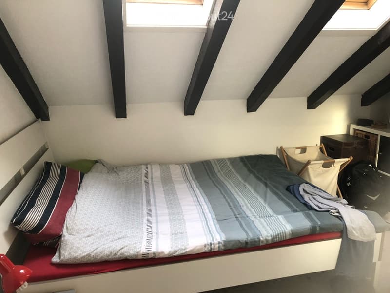 Cozy attic bedroom with slanted wooden beams, a single bed with striped bedding, a nightstand, and a small chair.