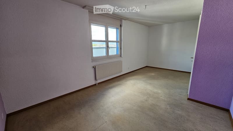 Empty room with light purple walls, a window with a view, and a radiator below the window. The floor is carpeted, and there are no furnishings.