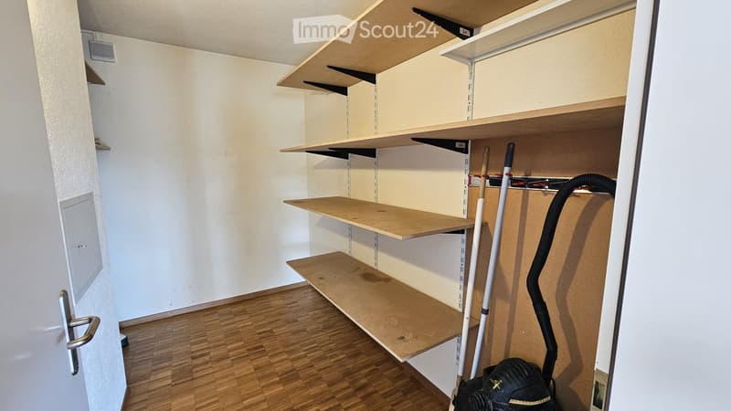 Empty closet with wooden shelves and a vacuum cleaner placed inside. The room has wooden flooring and white walls.
