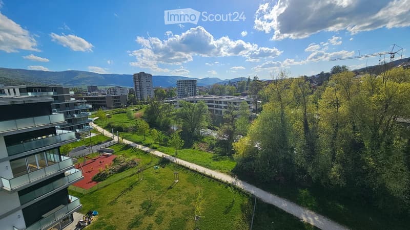Aerial view of a modern residential complex with green lawns, pathways, and surrounding buildings. The area includes a playground and seating areas.