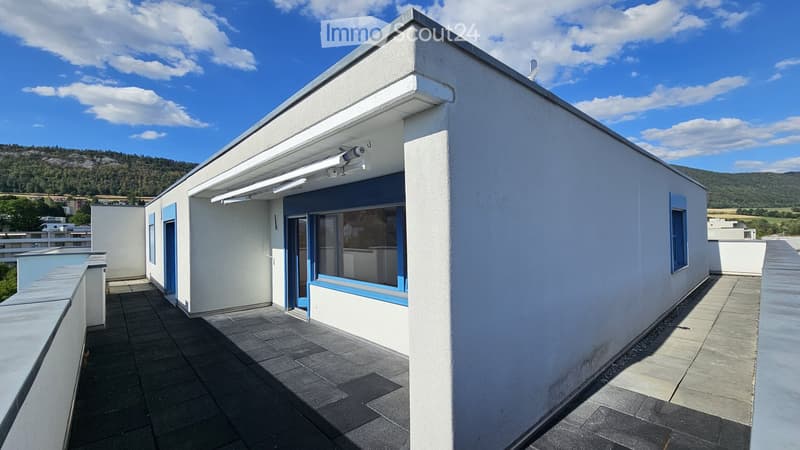 Modern rooftop terrace with grey tiled flooring, white walls, blue-framed windows, and a retractable awning. The background shows a scenic mountain view under a partly cloudy sky.