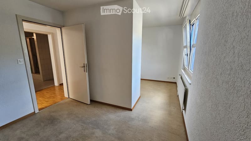 Empty hallway with carpeted floor, white walls, and a door leading to another room with wooden flooring.
