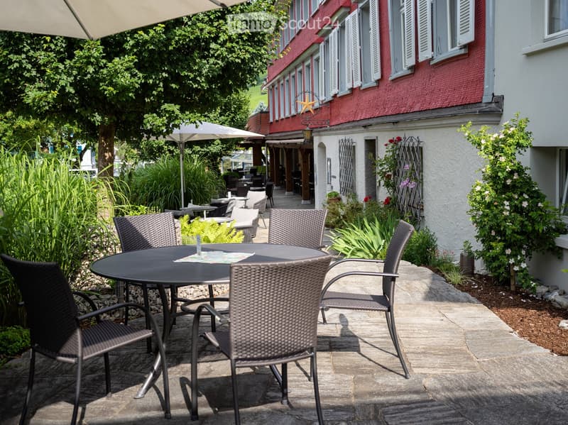 Outdoor seating area with a round table and several wicker chairs, surrounded by greenery and a building with red brick facade.