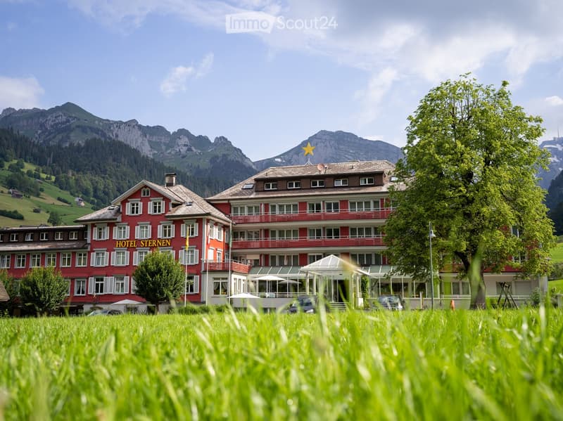 A large red hotel named "Hotel Sternen" with multiple stories, white-framed windows, balconies, and a prominent yellow star on the roof, surrounded by green grass and trees with mountain views in the background.