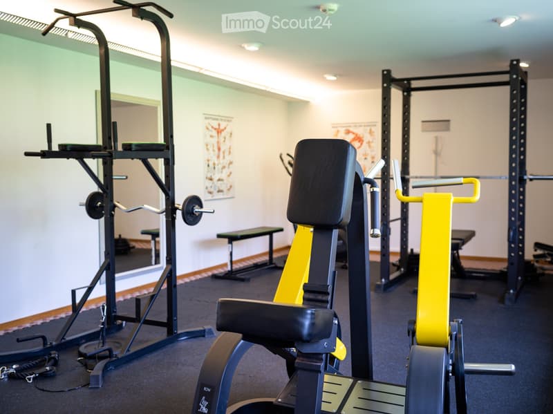 Well-lit gym room with various exercise equipment including weight machines, benches, and a mirror on the wall.
