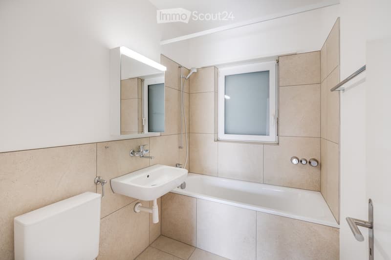 Bathroom with bathtub, sink, and shower area with showerhead. Beige tiles on the wall and tiled floor.