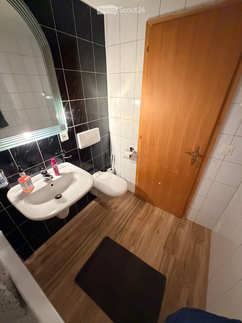 Modern bathroom with black and white tiled walls, a white sink with a chrome faucet, a toilet, a mirror with built-in lighting, and a wooden door.