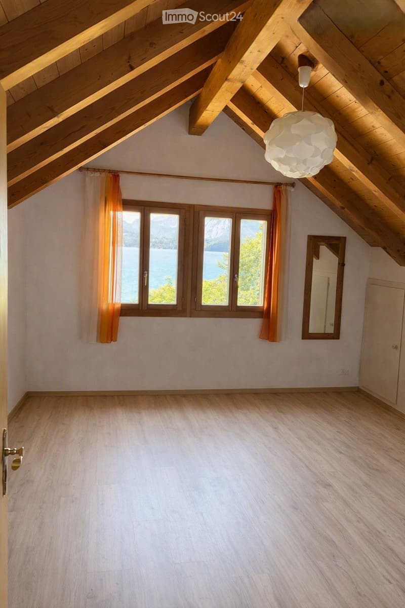 Empty attic room with wooden beams, large windows overlooking a lake, and a modern pendant light fixture.