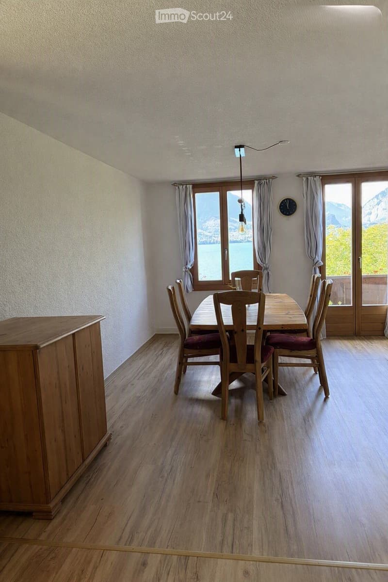 Bright dining room with wooden floor, large windows, and scenic mountain view. Features a wooden dining table with six chairs, a wall-mounted clock, and grey curtains.