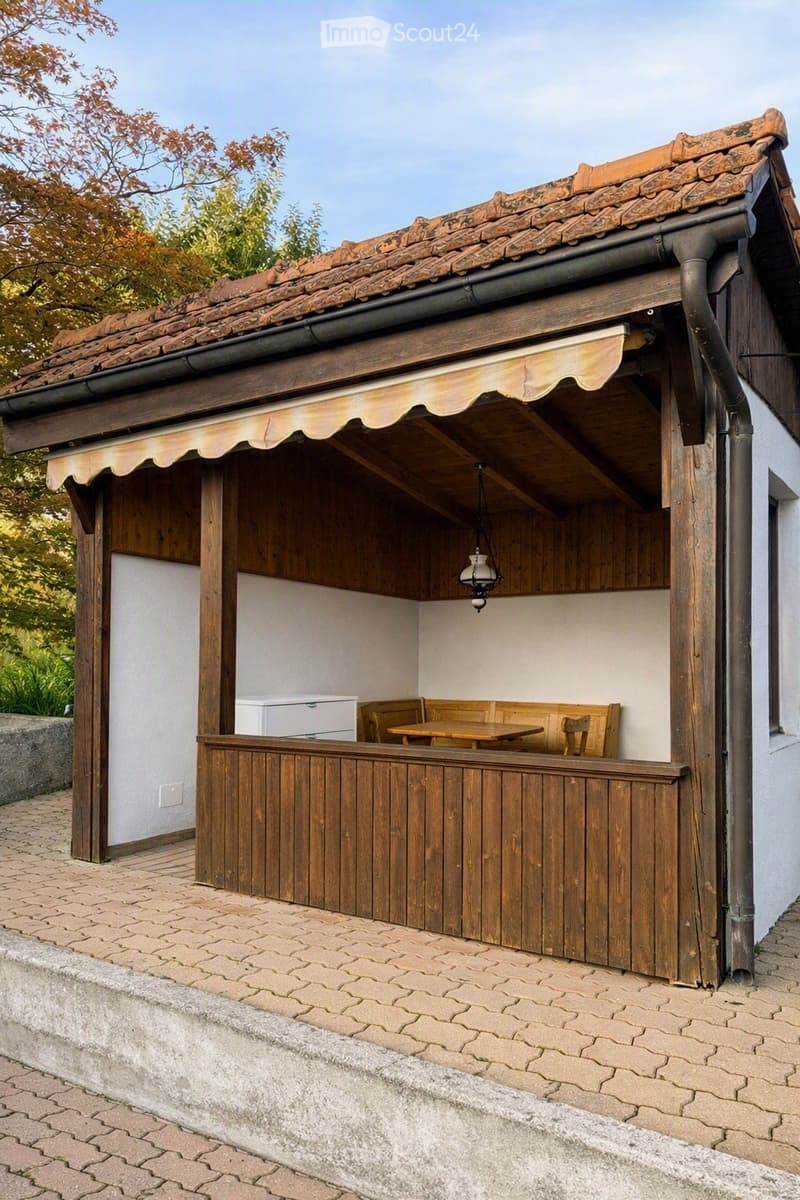 A wooden outdoor seating area with a table, chairs, and a bench, covered by a wooden awning with a tiled roof. The area is enclosed by wooden railings and has a small refrigerator in the background.
