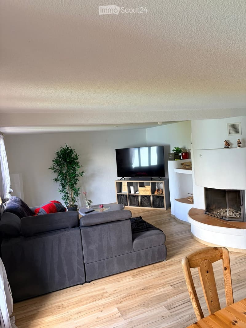 Modern living room with a grey sectional sofa, coffee table, TV on a shelf, fireplace, and a wooden dining chair near a small table.