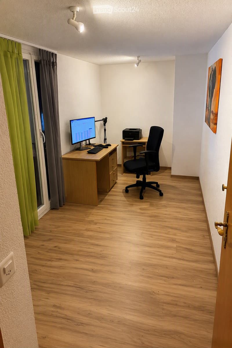 A well-lit room with wooden flooring, two desks, a computer setup, a black office chair, and a painting on the wall.