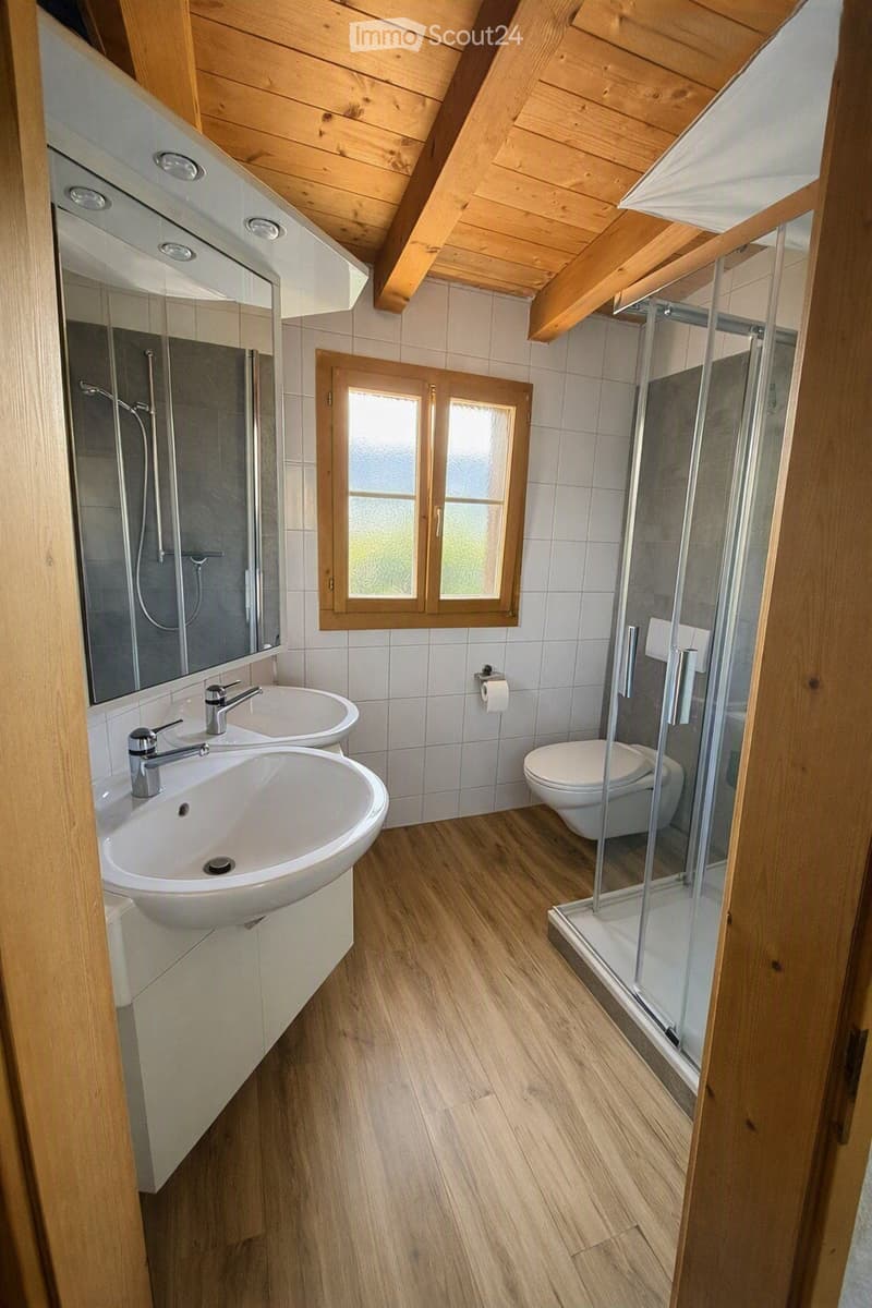 Modern bathroom with wooden flooring, double sink vanity, large mirror, toilet, and walk-in shower with glass enclosure.