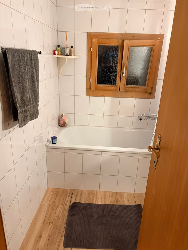 Bathroom with a white tiled wall, wooden floor, bathtub, towel, shelf, and a small mat in front of the tub.