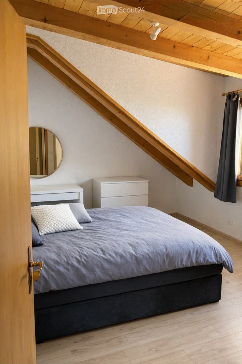 Cozy attic bedroom with wooden slanted ceilings, a neatly made bed with pillows and a duvet, a nightstand, a mirror, and a small drawer unit.