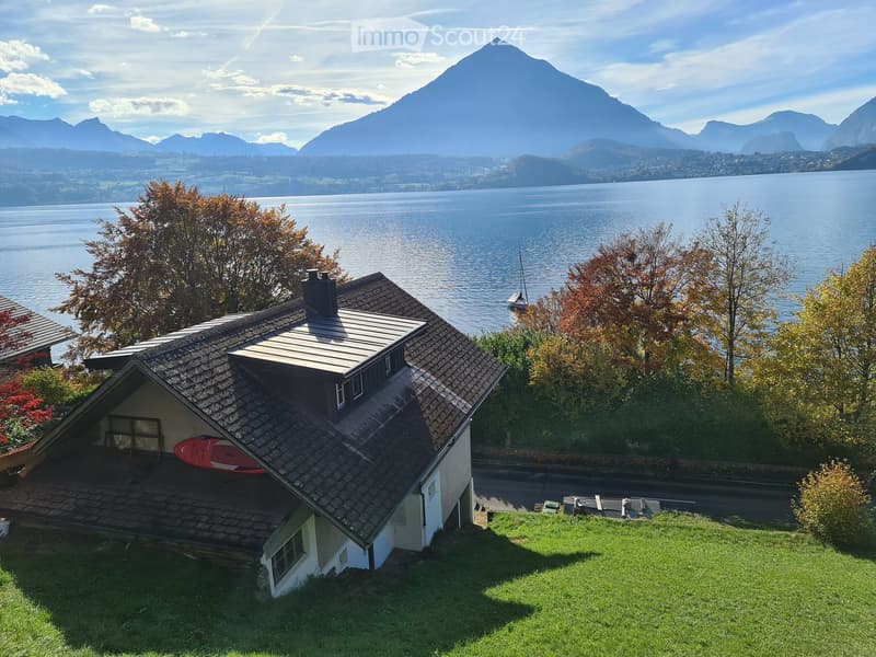 A two-story house with a tiled roof and a small garage, situated on a grassy slope overlooking a lake and mountains.