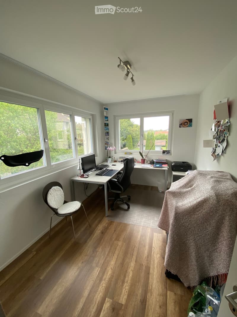 Bright, well-lit room with wooden flooring, large windows, and a desk setup including a computer, keyboard, and chair. A covered object is visible on the right side.