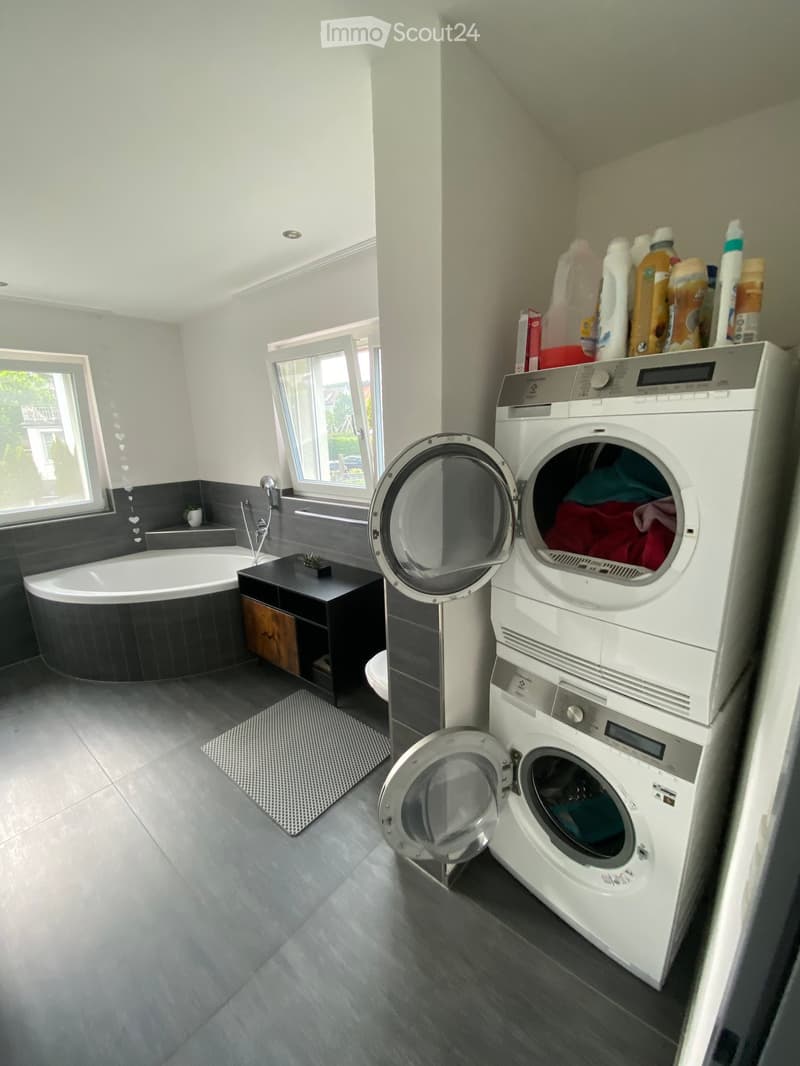 Modern bathroom with a bathtub, washing machine, dryer, small table, and a window letting in natural light.