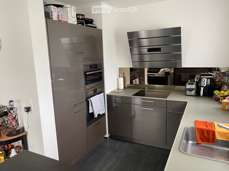 Modern kitchen with stainless steel appliances, grey cabinets, and a central island. Features include a refrigerator, oven, dishwasher, sink, and various small appliances on the counter.