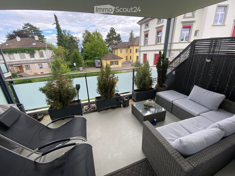 Balcony with wicker sofa, glass coffee table, lounge chairs, and potted plants, overlooking a residential area with trees and buildings.