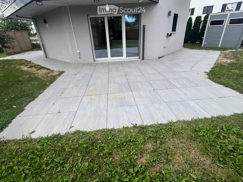 Outdoor patio area with large concrete tiles, a modern house entrance with large glass doors, and surrounding greenery.