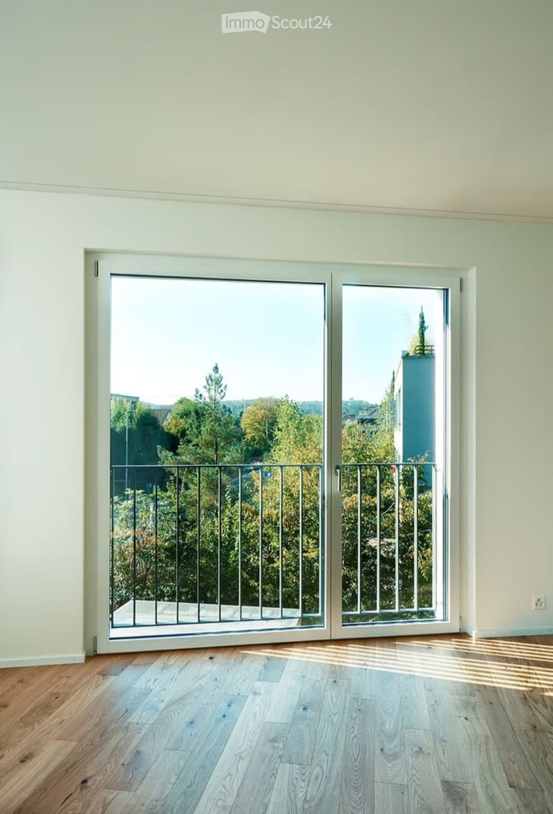 Empty room with light green walls, wooden flooring, and large sliding glass doors leading to a balcony with a metal railing and a view of trees and a building.