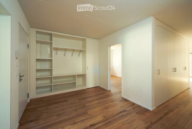 Empty room with wooden flooring, white walls, built-in closet with shelves and hanging space, and a door leading to another room.