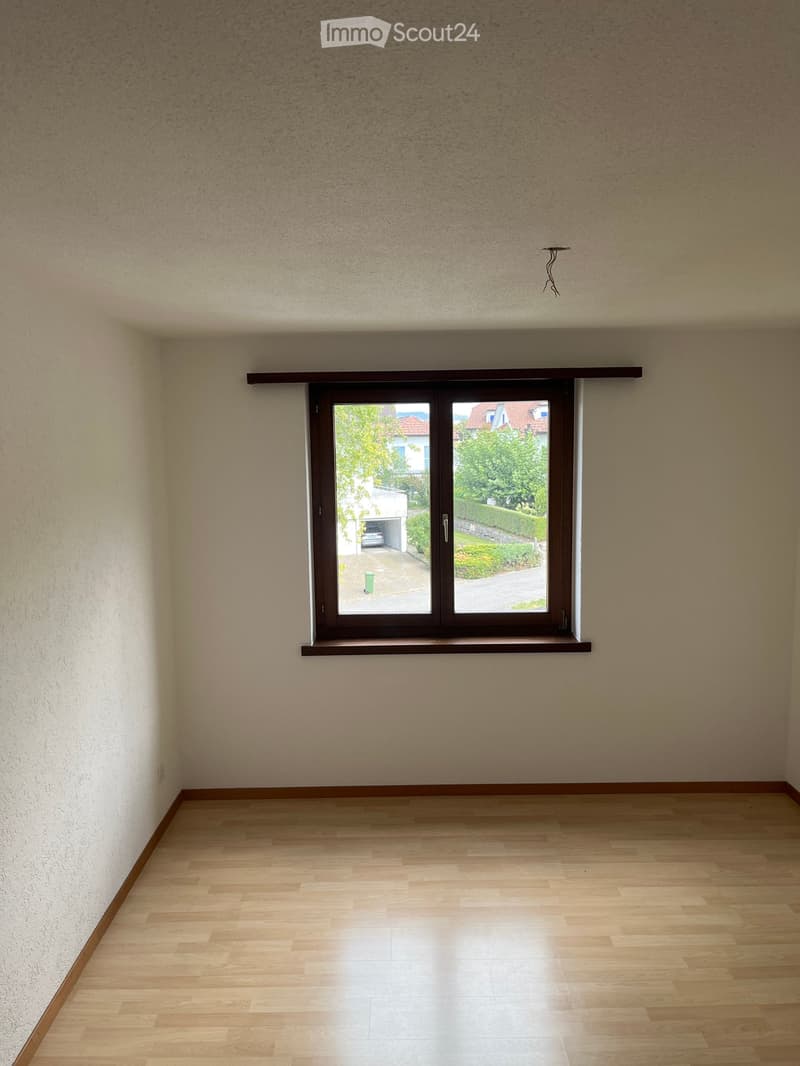 Empty room with light wooden flooring and white walls, featuring a large window with a brown frame and a small shelf above it.