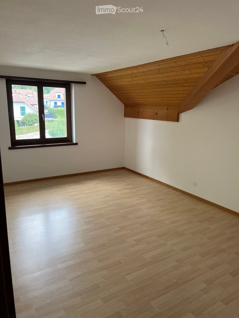 Empty room with wooden flooring, white walls, and a large window with a view of neighboring houses and greenery.