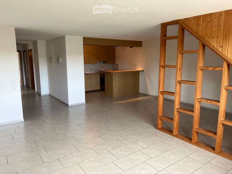 Empty room with tiled floor, white walls, wooden staircase, and adjacent kitchen area with cabinets and countertop.