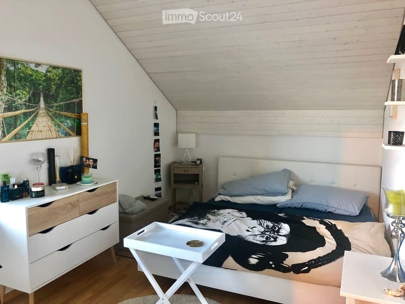 Cozy bedroom with a slanted wooden ceiling, a white upholstered bed with blue pillows, a nightstand with a lamp, a white dresser with drawers, a small table, and a large picture frame on the wall.