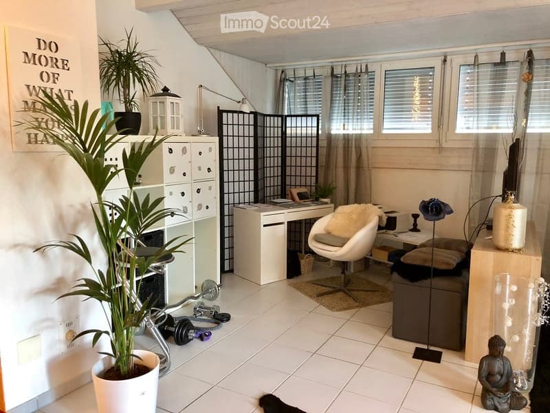 A well-lit room with a white tiled floor, a large potted plant, a white desk with a chair, a grey sofa with cushions, a floor lamp, a decorative shelf, and a small fitness area with weights.