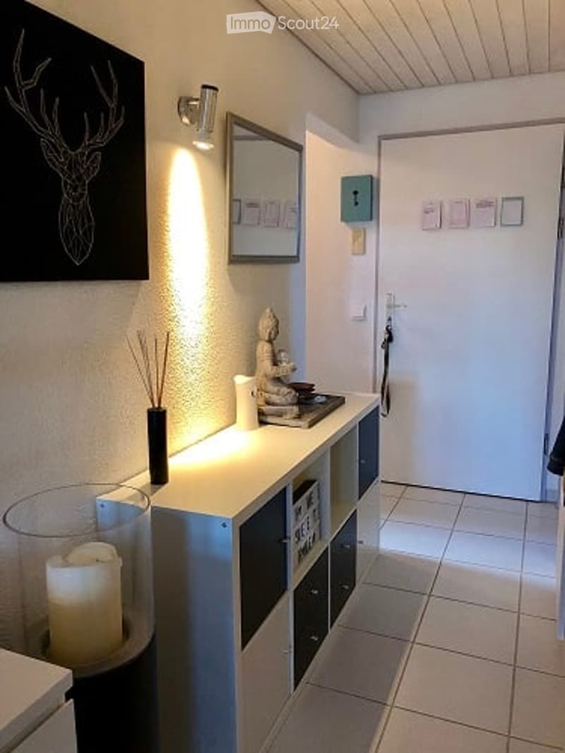 Modern hallway with white walls, tiled floor, built-in storage unit with shelves and drawers, decorative items including a candle, vase, and small statue, illuminated by wall-mounted lighting.