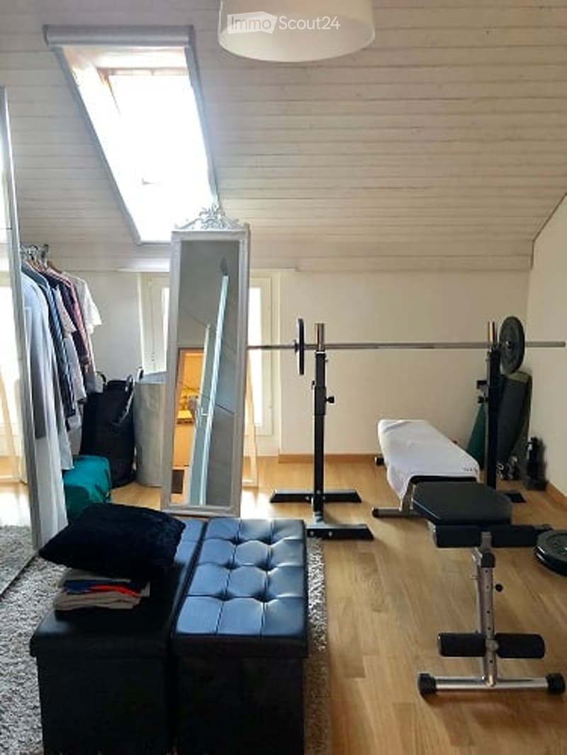 Well-lit room with wooden flooring, a large mirror, a barbell setup, a bench, and a stool with folded clothes.