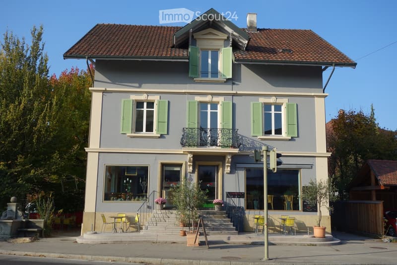 Two-story building with grey facade, green shutters, and a balcony on the second floor. The ground floor has large windows and a door leading to a small outdoor seating area with tables and chairs.