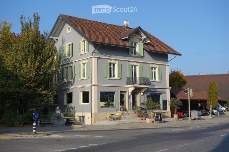 Two-story building with grey facade, green shutters, and a red-tiled roof. Features a small balcony, outdoor seating area, and a storefront with large windows.