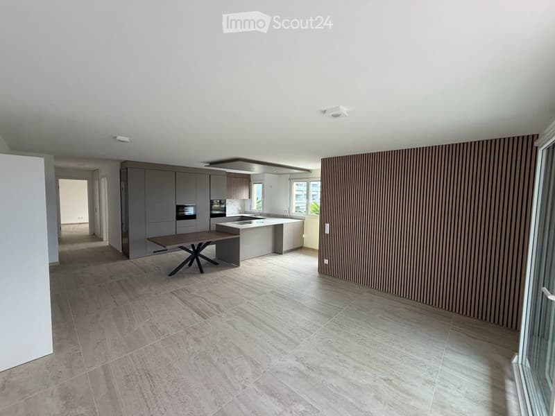 Modern kitchen and dining area with grey cabinets, a central island, a wooden dining table, and a sleek oven. The space features large windows and a contemporary design with a mix of wood and metal finishes.