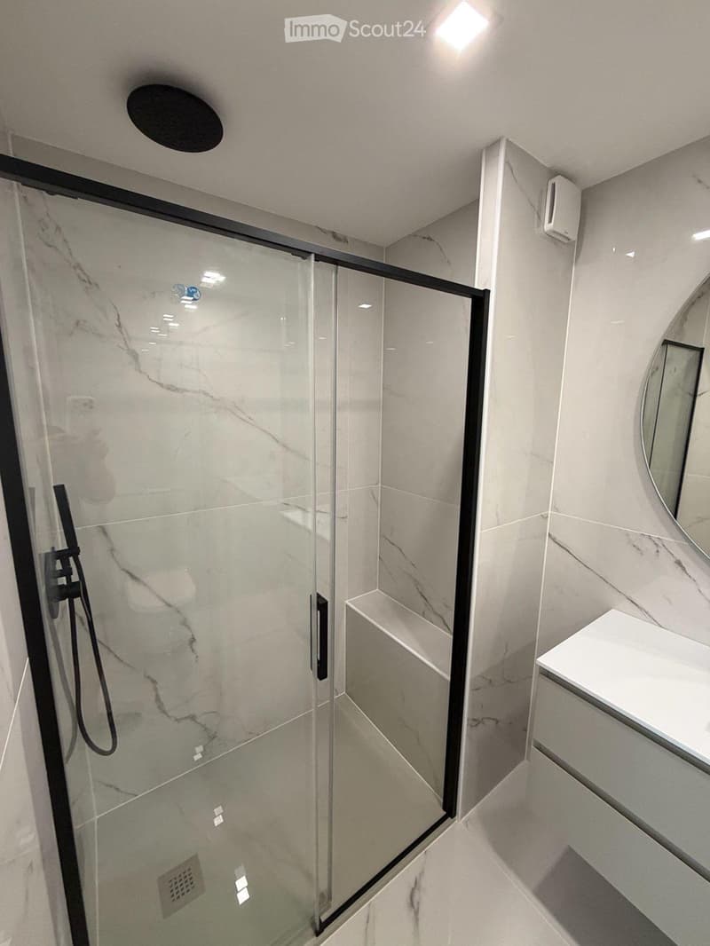 Modern bathroom with a glass-enclosed shower stall featuring marble tiles, a white sink with a mirror, and sleek black fixtures.