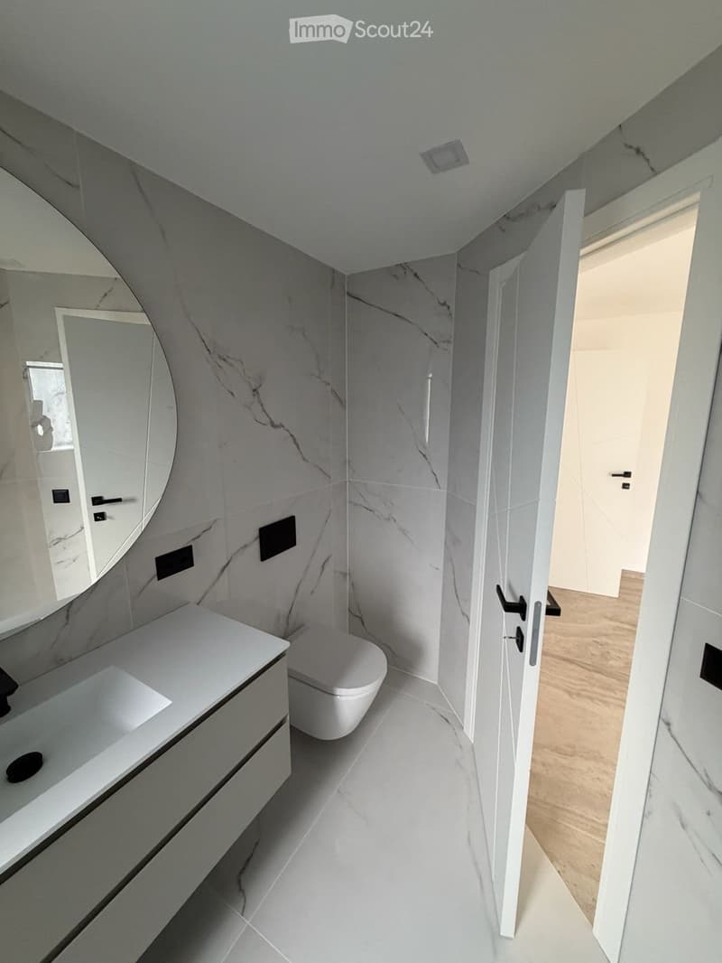 Modern bathroom with white marble tiles, a floating sink, a toilet, and a large round mirror.
