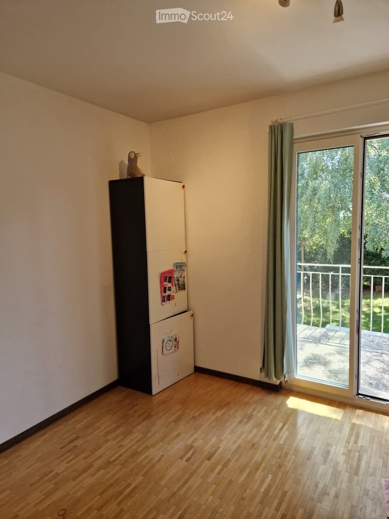 A small, unfurnished room with wooden flooring, a refrigerator, and a sliding glass door leading to a balcony.