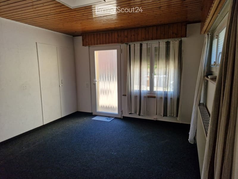 Empty room with wooden ceiling, carpeted floor, white walls, and a glass door leading outside. The room has a radiator and a small shelf visible.