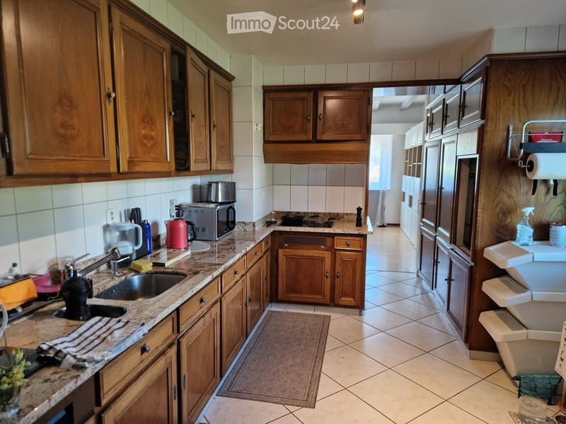 A well-equipped kitchen with wooden cabinets, granite countertops, a sink, microwave, toaster, and refrigerator. The space is clean and organized with tiled floors and walls.