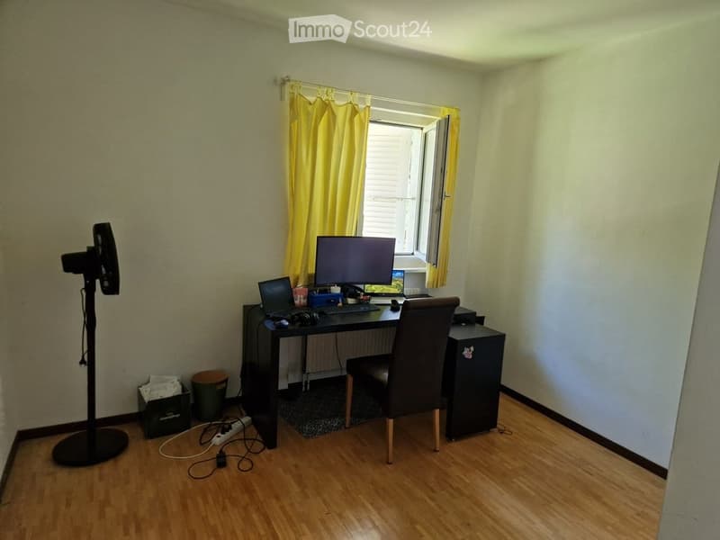 A small, well-lit room with wooden flooring, a desk setup including a laptop, monitor, and chair, a standing fan, and a radiator under the window.