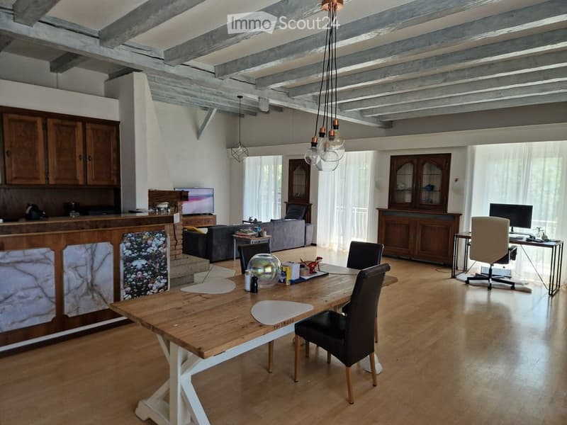 Open-plan living and dining area with exposed wooden beams, a large wooden dining table with chairs, a sofa, a television, and a fireplace. Wooden cabinets and pendant lights are also visible.