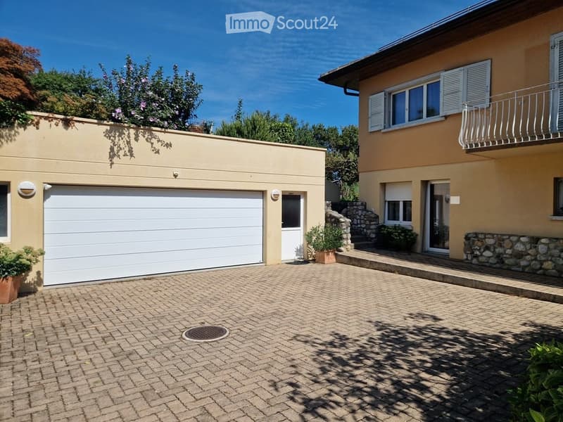 Two-story house with a garage, beige exterior, white shutters, and a brick driveway.