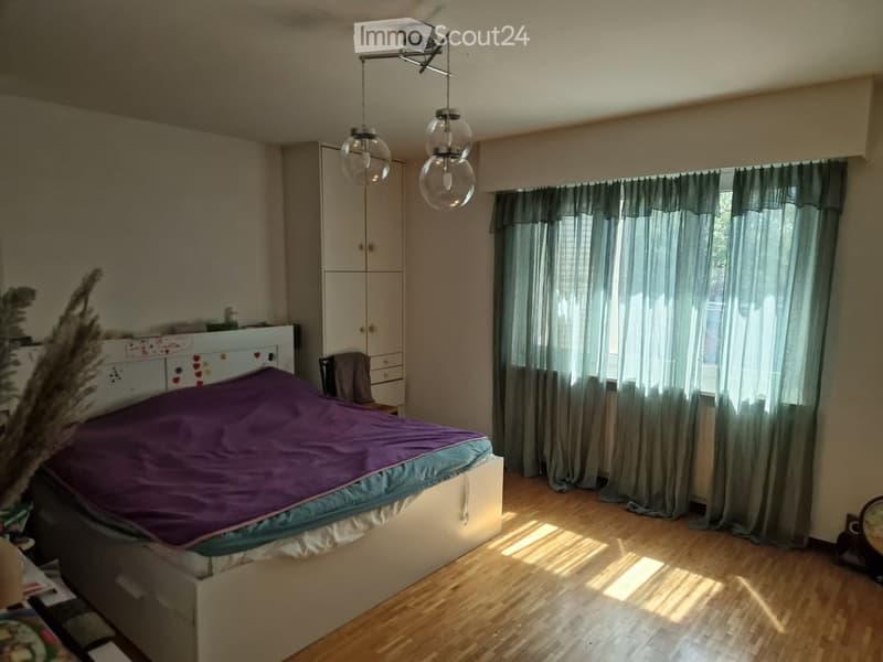 Bedroom with a purple duvet on the bed, a white wardrobe, a chair, and a window with grey curtains. The room has wooden flooring and a modern pendant light fixture.