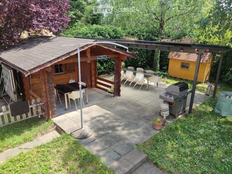 Outdoor patio area with a wooden cabin, dining table and chairs, grill, and small shed surrounded by greenery.